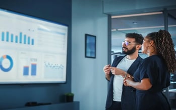 A man and a woman looking at a big screen showing a dashboard